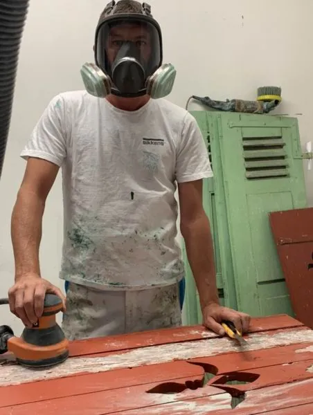 Ponçage d’un volet à la machine, l’artisan porte un masque